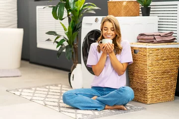 Abwaschbare Fototapete Beautiful smiling woman, housewife holding cup with drink  sitting in lotus position in modern laundry room with washing machine on background, copy space © Maria Vitkovska