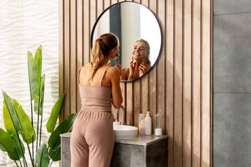 Back view of smiling cute woman using jade roller doing face massage looking in mirror in modern...