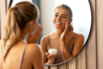 Beautiful smiling woman applying moisturizer cream on her face looking in mirror. Skin care,...