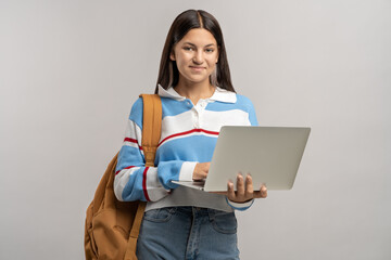 Fototapeta premium Pleased teenage school girl with laptop. Happy satisfied excited young student girl smiling with backpack on shoulder, holding laptop computer in hands, preparing essay, term paper, studying online.