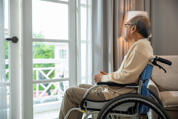 Asian senior man disabled sitting alone in wheelchair looking through window at hospital, lonely elder thoughtful sad old man look outside windows in bedroom at retirement home, feel depressed lonely