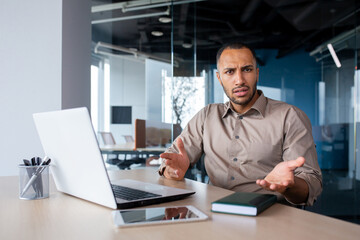 Dissatisfied hispanic male office worker sitting at desk indoors at desk, working on laptop, spreading hands surprised indignant, looking at camera.