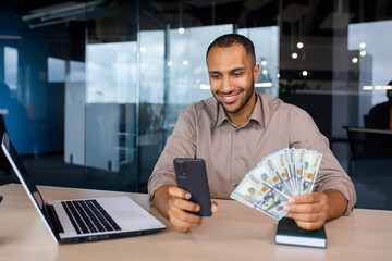 Successful businessman sitting at the table in the office in front of the laptop, holding the phone...