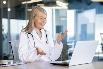 Mature experienced female doctor talking remotely using video call, gray haired woman hospital worker consulting patients sitting inside modern clinic office in white medical coat.