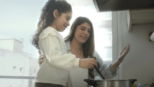 Little girl stirring boiling food in cooking pot and talking with joyful mother while preparing dinner together in domestic kitchen