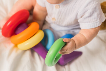 Close-up baby playing with colorful rainbow toy pyramid on white sunny bedroom. Toys for little kids. Child with educational toy.
