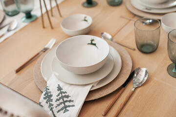 minimal scandinavian trendy Christmas table setting of white plates with green reindeer print, napkin and wooden table and decor cutlery - obrazy, fototapety, plakaty