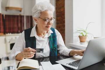 Happy smiling senior female with gray hair in formal wear sitting at kitchen table holding plastic card in hands doing online payment using laptop and banking application, paying utility bill