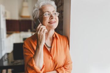 Portrait of happy smiling elderly female with gray hair in stylish shirt and glasses talking on phone leaning against wall, enjoying nice conversation with best friend living abroad