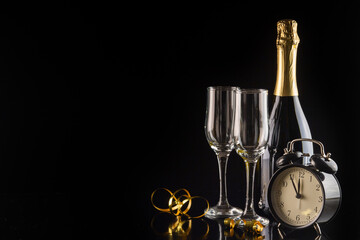Alarm clock, champagne bottle and glasses on black background with copy space. New Year and Christmas celebration concept.
