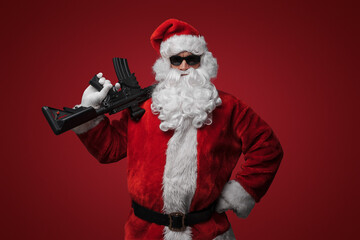 A man dressed as Santa Claus, wearing sleek black sunglasses, poses with toy machine guns against a...