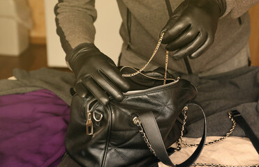 Robber in black outfit and gloves see in opened stolen women bag. The thief takes out golden...
