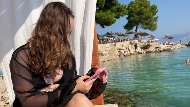 Internet wi-fi good network connection social networks a young girl sits on a VIP lounger with Curtains near the sea ocean people swim in the background Albania fir trees and mountains. rocks