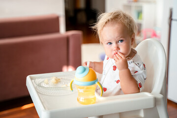 A little girl child sits on a high chair in the kitchen and eats, complementary food