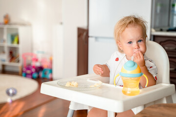 A little girl child sits on a high chair in the kitchen and eats, complementary food