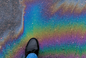 A man's foot steps on a rainbow stain of gasoline on the asphalt. Gasoline stains pollute the...
