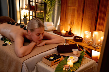 Obraz na płótnie Canvas Hot stone massage at spa salon in luxury resort with warm candle light, blissful woman customer enjoying spa basalt stone massage glide over body with soothing warmth. Quiescent