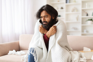 Sick indian guy sitting on couch covered in blanket