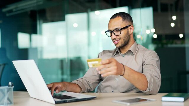 Handsome happy business man making bank payment, holding credit card sitting at computer in modern office. male entrepreneur at desk to pay for bills, making purchase online. Golden customer order