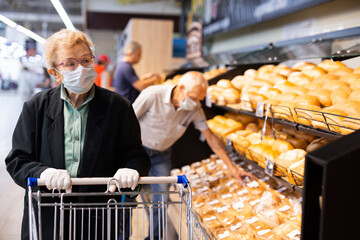 Older woman with glasses chooses buns and bread in supermarket bakery