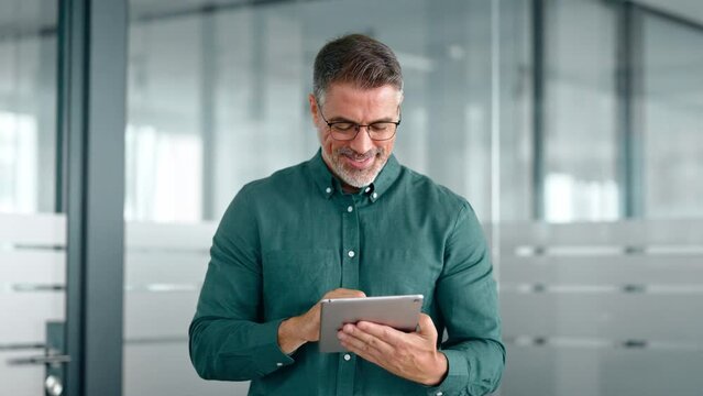 Smiling busy middle aged business man investor using tablet computer, mature older businessman executive manager looking at tab satisfied with finance trading market standing in office.