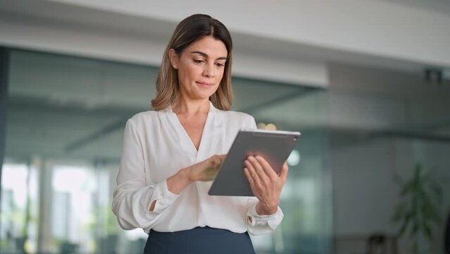 Mature 40 years old smiling busy professional latin business woman manager executive or entrepreneur holding tab using digital tablet standing in office working on corporate device.