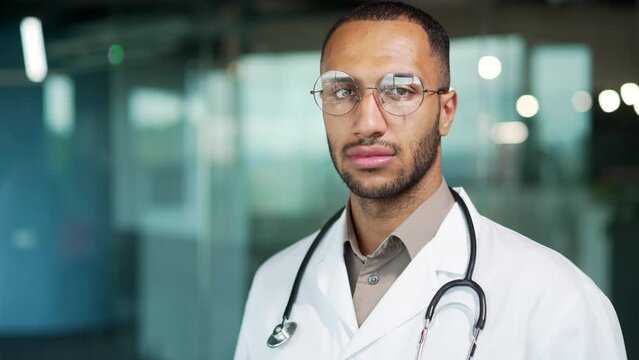 Portrait young serious confident focused doctor looking at camera while standing in modern office clinic close up. Handsome mixed race physician in glasses and white coat posing Head shot doc Closeup