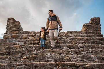 Photographer man and his fashionable little son sightseeing together in a castle walking down stairs hand in hand while looking at the surroundings, traveling and photographing a small town - obrazy, fototapety, plakaty