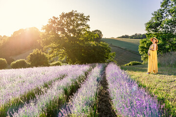 A pregnant woman in a yellow dress stands on the edge of a field of blooming lavender in the rays of the setting sun