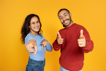 Positive young man and woman pointing at camera on yellow