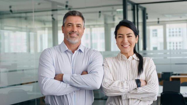 Happy confident professional mature Latin business man and Asian business woman corporate leaders managers standing in office, two diverse executives team posing arms crossed, portrait.