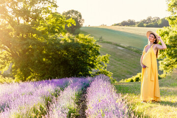 A pregnant woman in a yellow dress stands on the edge of a field of blooming lavender in the rays of the setting sun