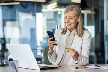 Senior businesswoman working in office at desk, using mobile phone and credit card. - Powered by Adobe