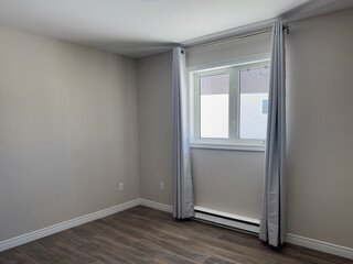 An empty master bedroom with a white ceiling, beige painted walls and dark brown wooden hardwood floor in the interior of a house. Grey curtains are hanging from a large closed glass window.