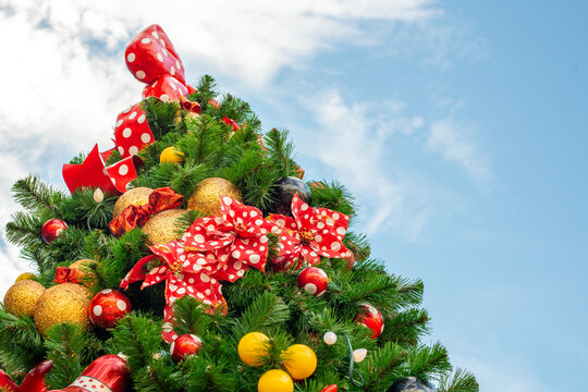 A tall fir tree decorated with glass balls, toys, ornaments, Christmas lights, glitter, red polka dot bows,  bobbles, and baubles outdoors under a blue sky. There's a red ribbon looped at the top.
