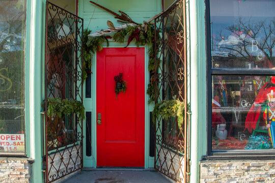 The exterior facade of a vintage shop or store with an orange color wooden door decorated with a Christmas garland. The items in the large glass windows are antique trinkets and toys. 