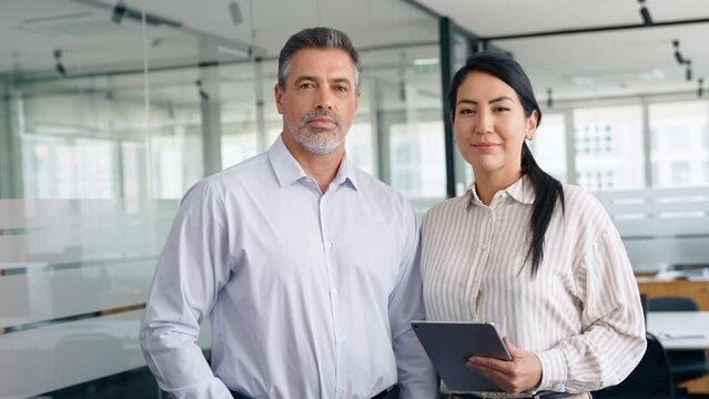Happy confident professional mature Latin business man and Asian business woman corporate leaders managers standing in office, two diverse colleagues executives team together, portrait.