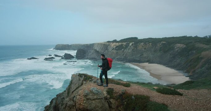 Wide angle establishing shot of male hiker with red backpack and trekking poles stand on edge of cliff overlooking ocean landscape. Man hiking alone on epic west coast trail