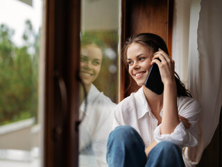 Young woman sitting on a wooden window sill by the window with a phone in hand smiling and looking...