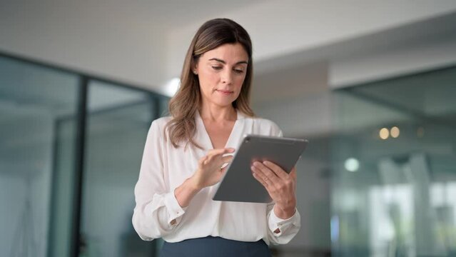 Middle aged 45 years old busy professional business woman company manager sales executive holding fintech tab using digital tablet computer standing in office working on corporate pad.