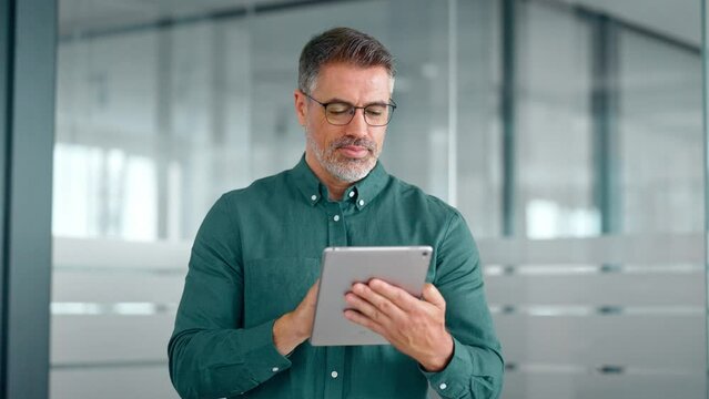 Busy middle aged business man investor using tablet computer, mature older businessman executive manager looking at tab satisfied with finance trading market working standing in office.