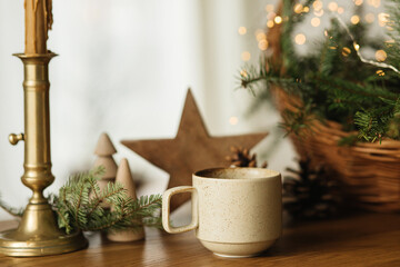 Stylish cup of warm tea, fir branches in basket, wooden trees and star, pine cones and lights on table. Modern christmas rustic eco friendly decor, scandinavian winter hygge. Atmospheric christmas
