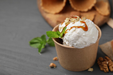 Tasty ice cream with caramel sauce, mint and nuts in paper cup on grey wooden table, closeup. Space for text