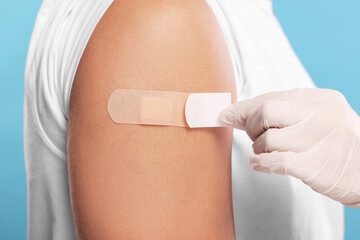 Doctor sticking plaster on man's arm after vaccination against light blue background, closeup