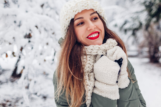 Close up portrait of happy young woman in snowy winter park wearing warm knitted hat scarf and red festive lipstick.