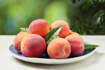 Many whole fresh ripe peaches and green leaves in plate on white table against blurred background, closeup