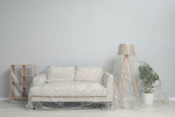 Sofa, shelving unit, lamp and houseplant covered with plastic film near light wall indoors. Space...
