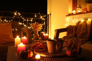 Rattan furniture, cups, fairy lights, burning candles and other autumn decor on outdoor terrace at...