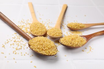Spoons with raw bulgur on white tiled table, closeup