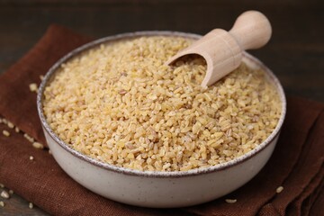 Bowl and scoop with raw bulgur on table, closeup